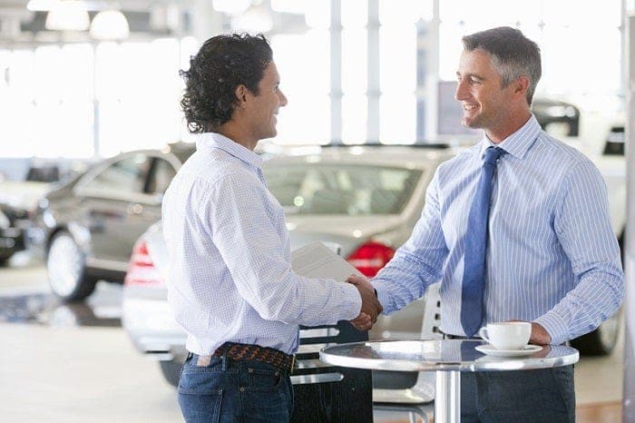 A smiling salesman and customer shaking hands inside a bright car dealership showroom.