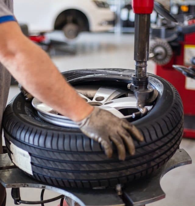 Mechanic's gloved hand mounting a black tire onto a silver rim using a machine.